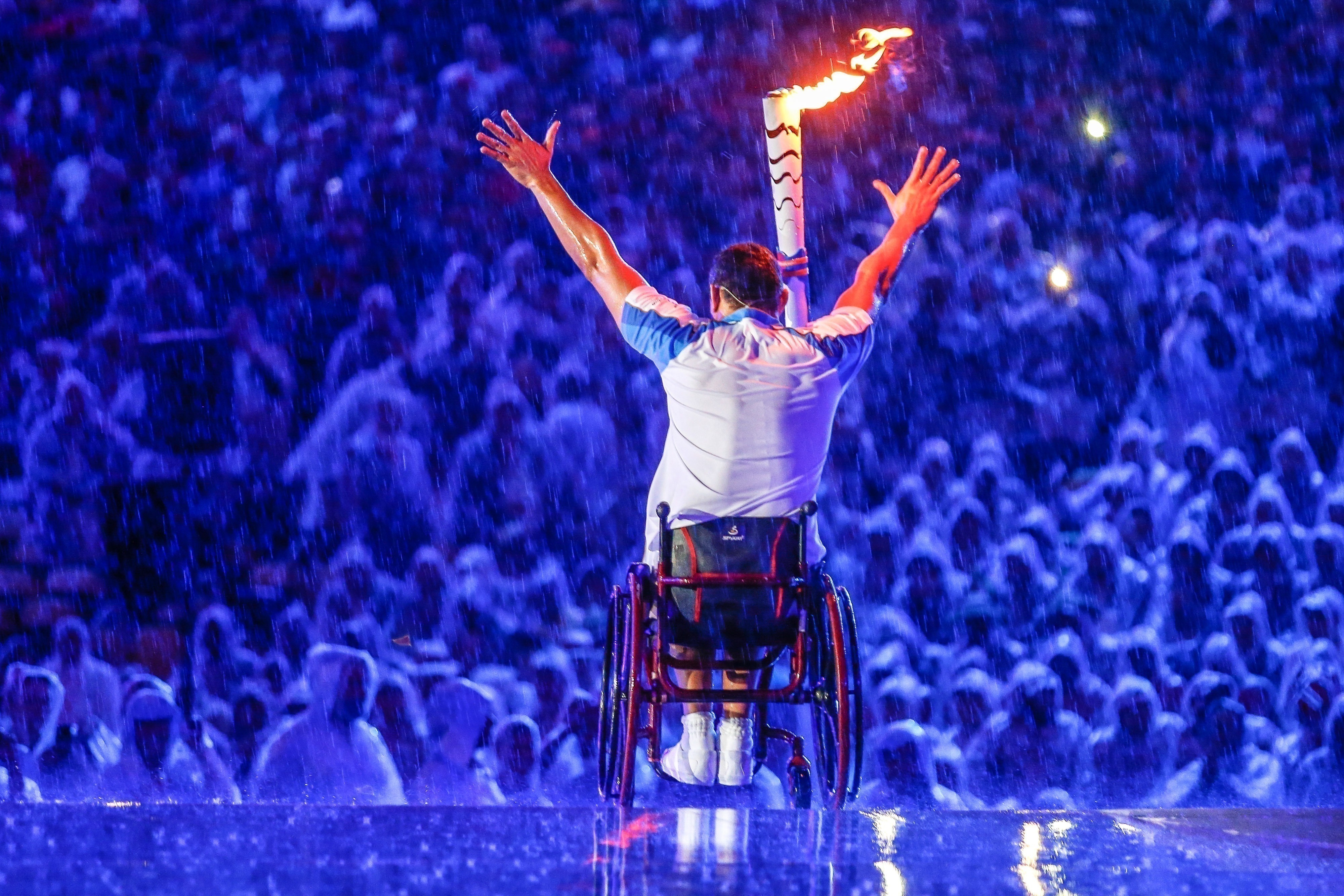 Clodoaldo Silva acena de sua cadeira, com pira olímpica acesa, para platéia embaixo de chuva.